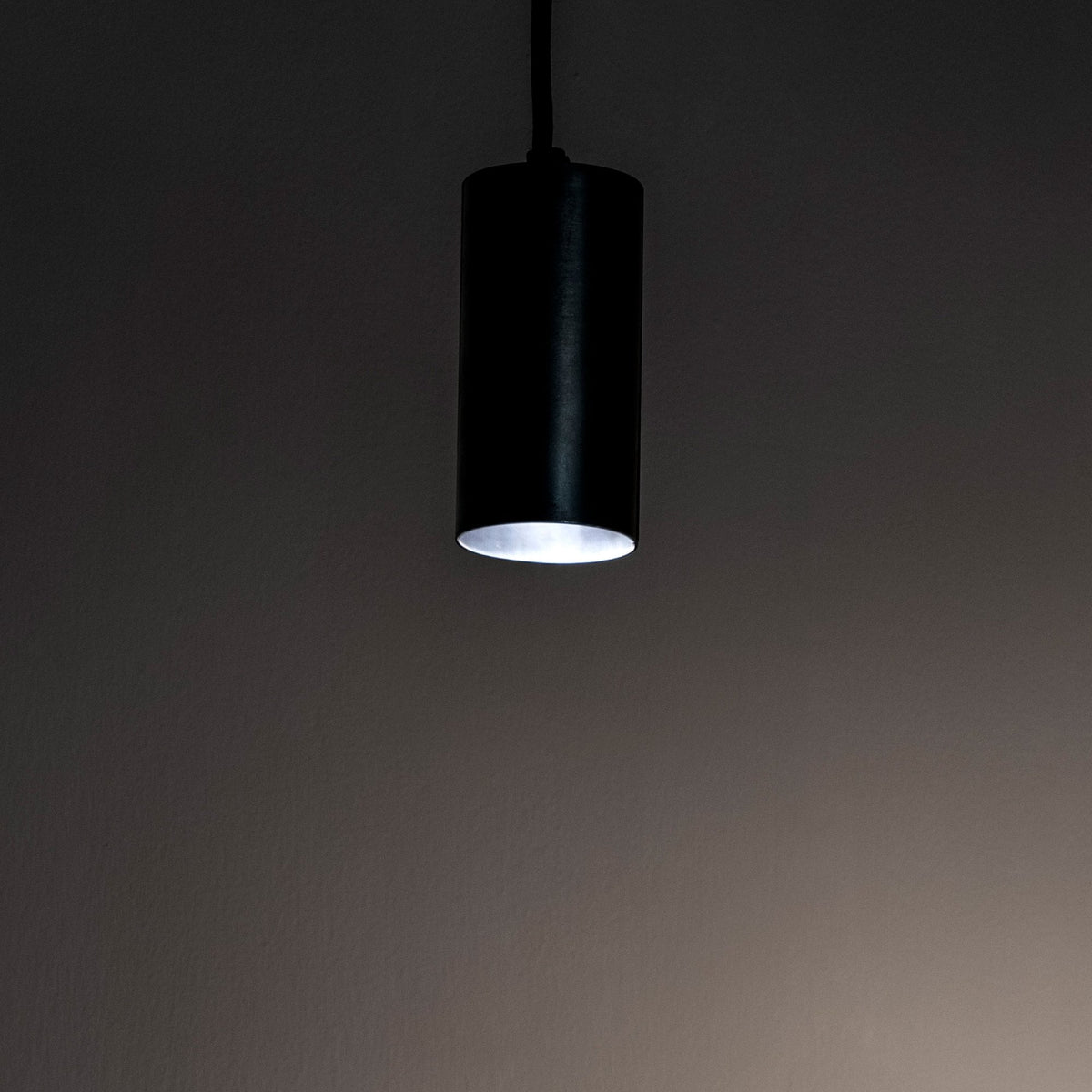 A minimalist cylindrical pendant light emitting a soft white glow in a dim room.