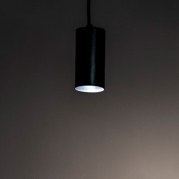 A minimalist cylindrical pendant light emitting a soft white glow in a dim room.