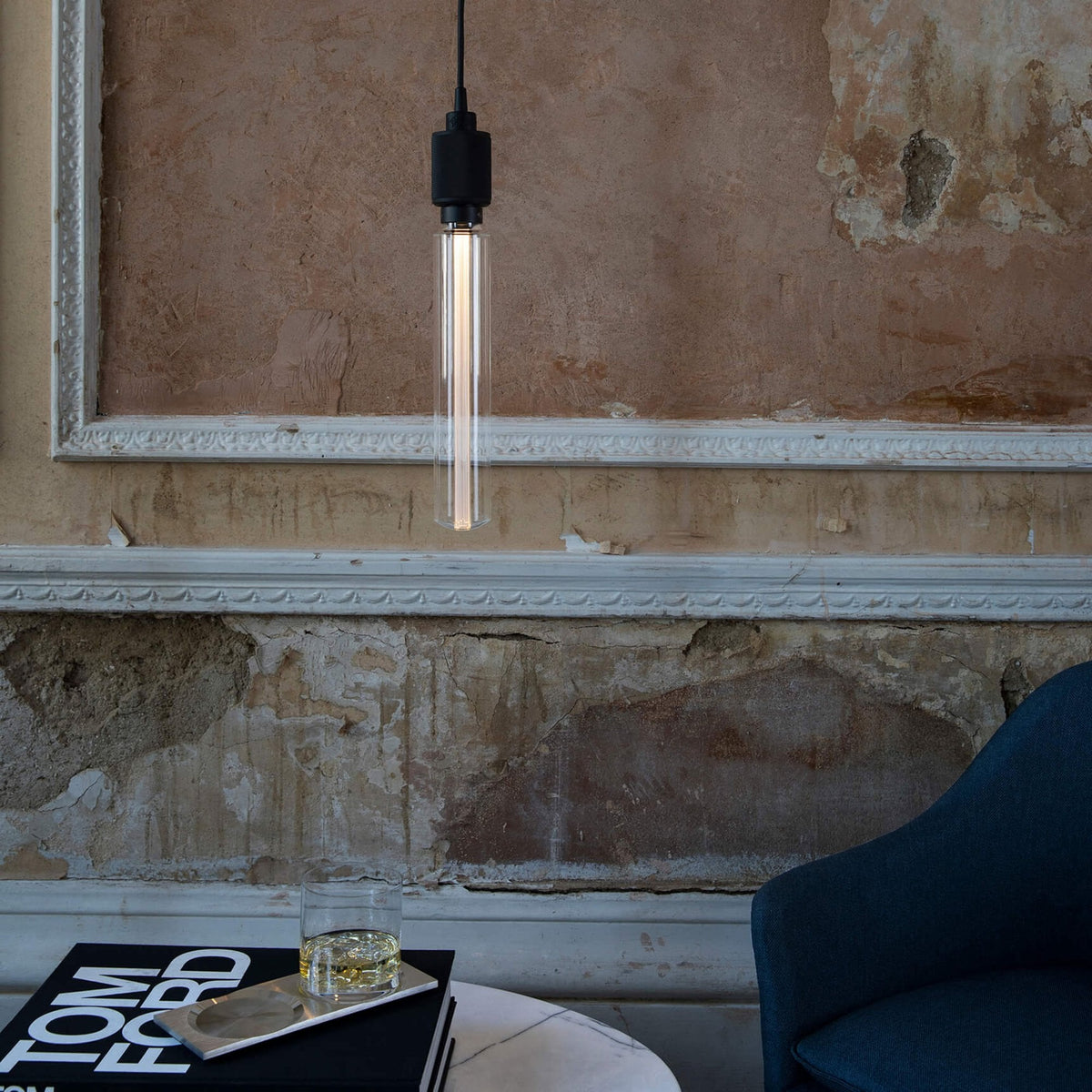 A modern cylindrical pendant light hangs in a room with distressed walls, next to a book and a drink.