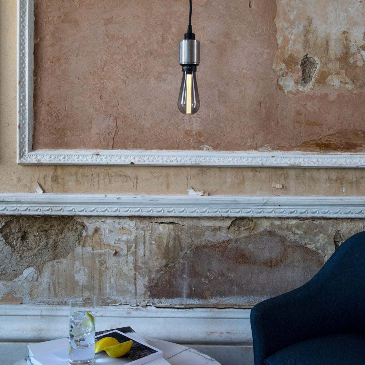 A modern lightbulb hangs from the ceiling against a distressed wall with decorative molding. A drink and lemons sit on a table.