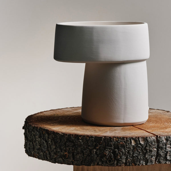 A minimalist white ceramic table lamp with a wide, flat top on a raw wooden table slice.