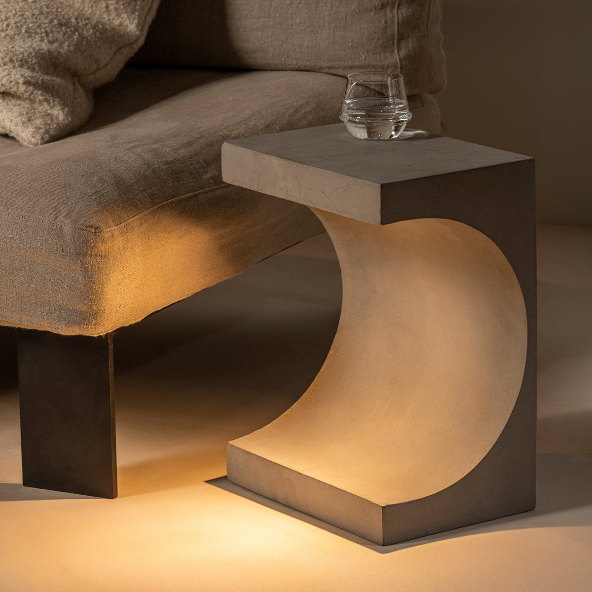 Concrete table lamp with a curved cut-out emitting a warm glow next to a sofa.