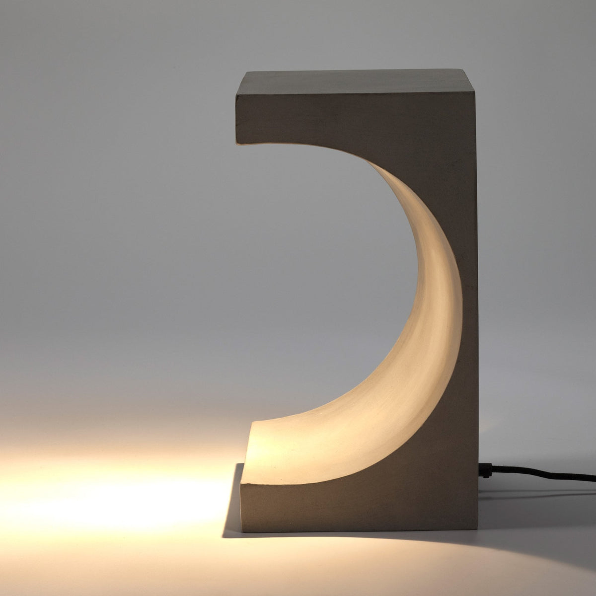 Concrete table lamp with a curved cut-out emitting a warm glow onto the surface.