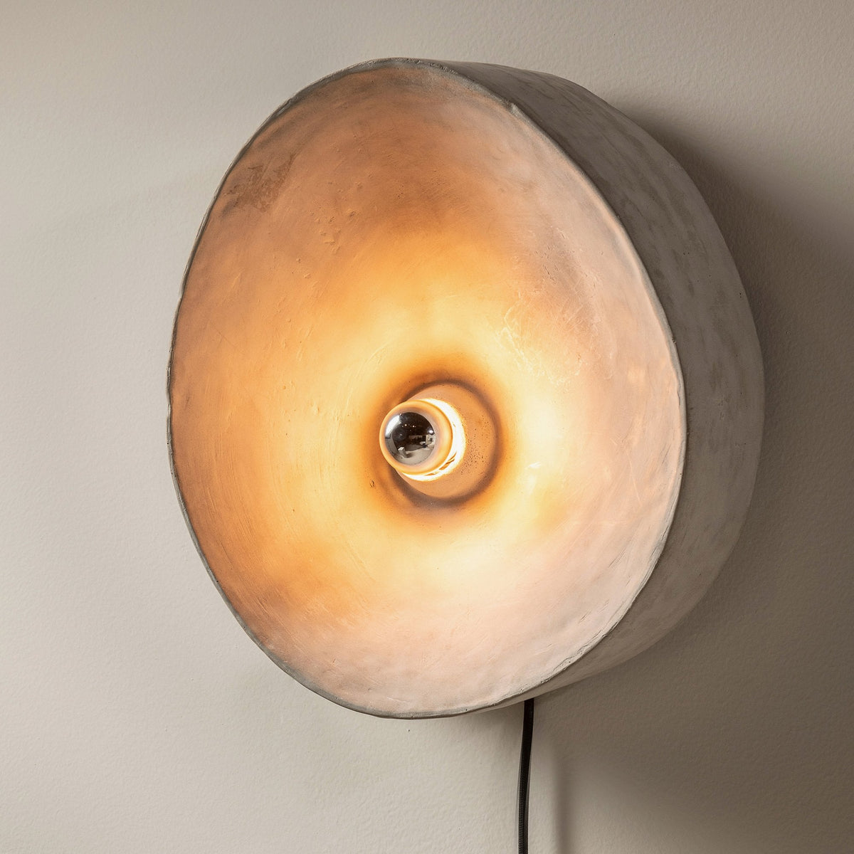 A close-up of a circular grey wall light with a warm, glowing bulb facing outwards.