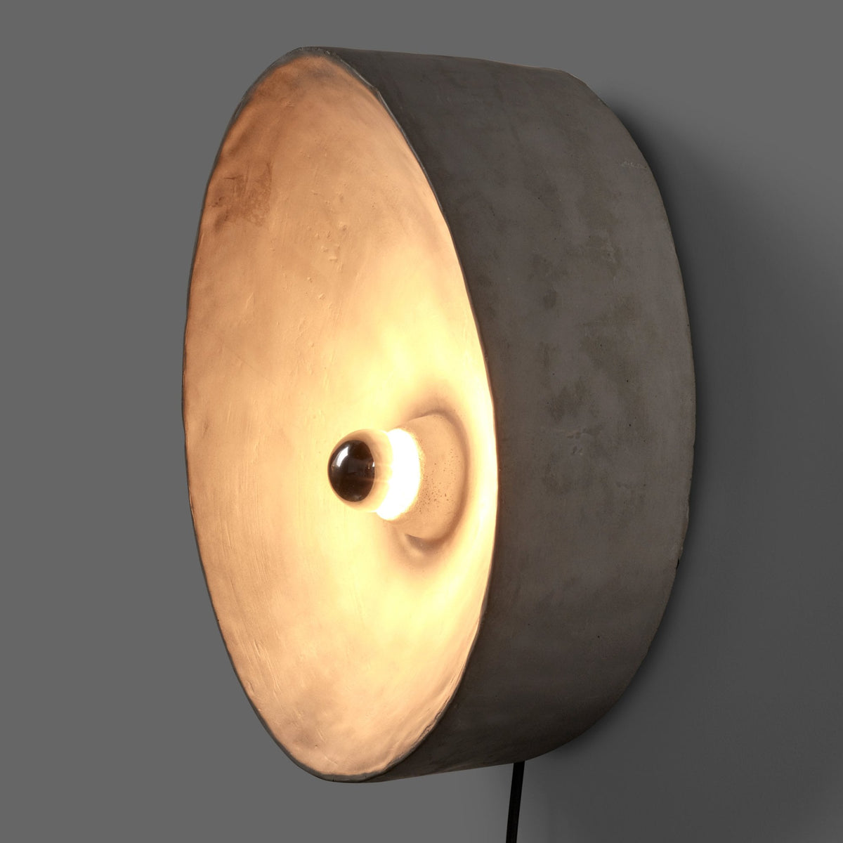 A side view of a grey circular wall light emitting a warm glow from its central bulb.