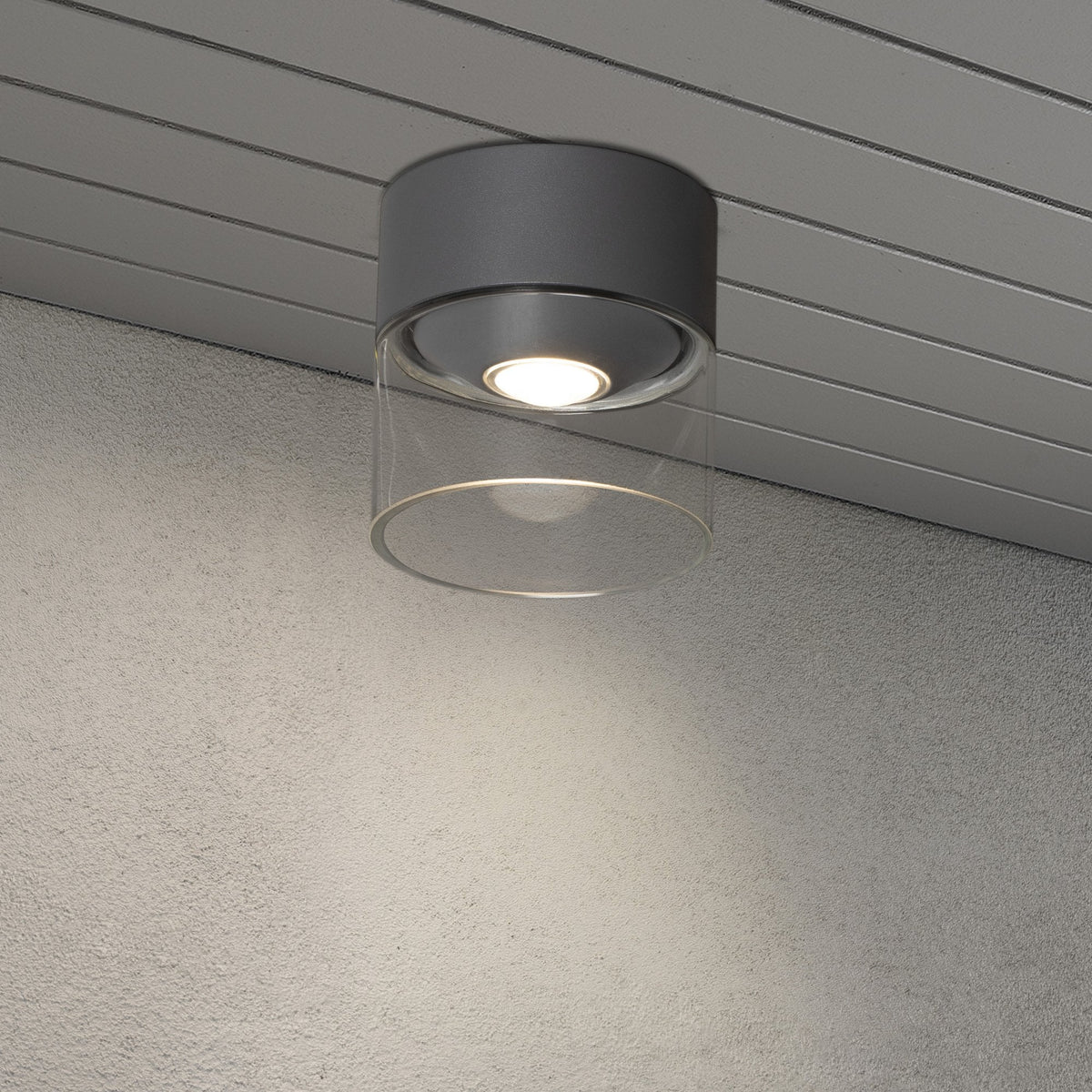 A modern grey and clear cylindrical outdoor ceiling light illuminating a concrete wall.