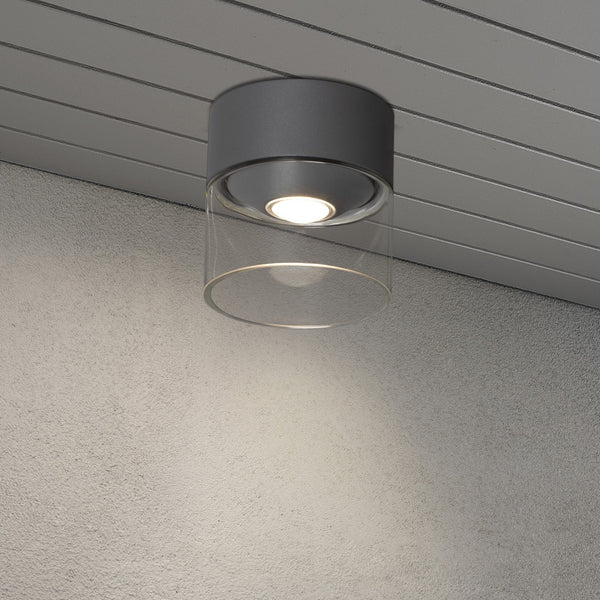 A modern grey and clear cylindrical outdoor ceiling light illuminating a concrete wall.