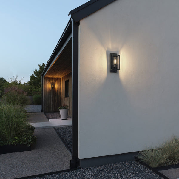 An outdoor wall light illuminating the side of a modern house at dusk.