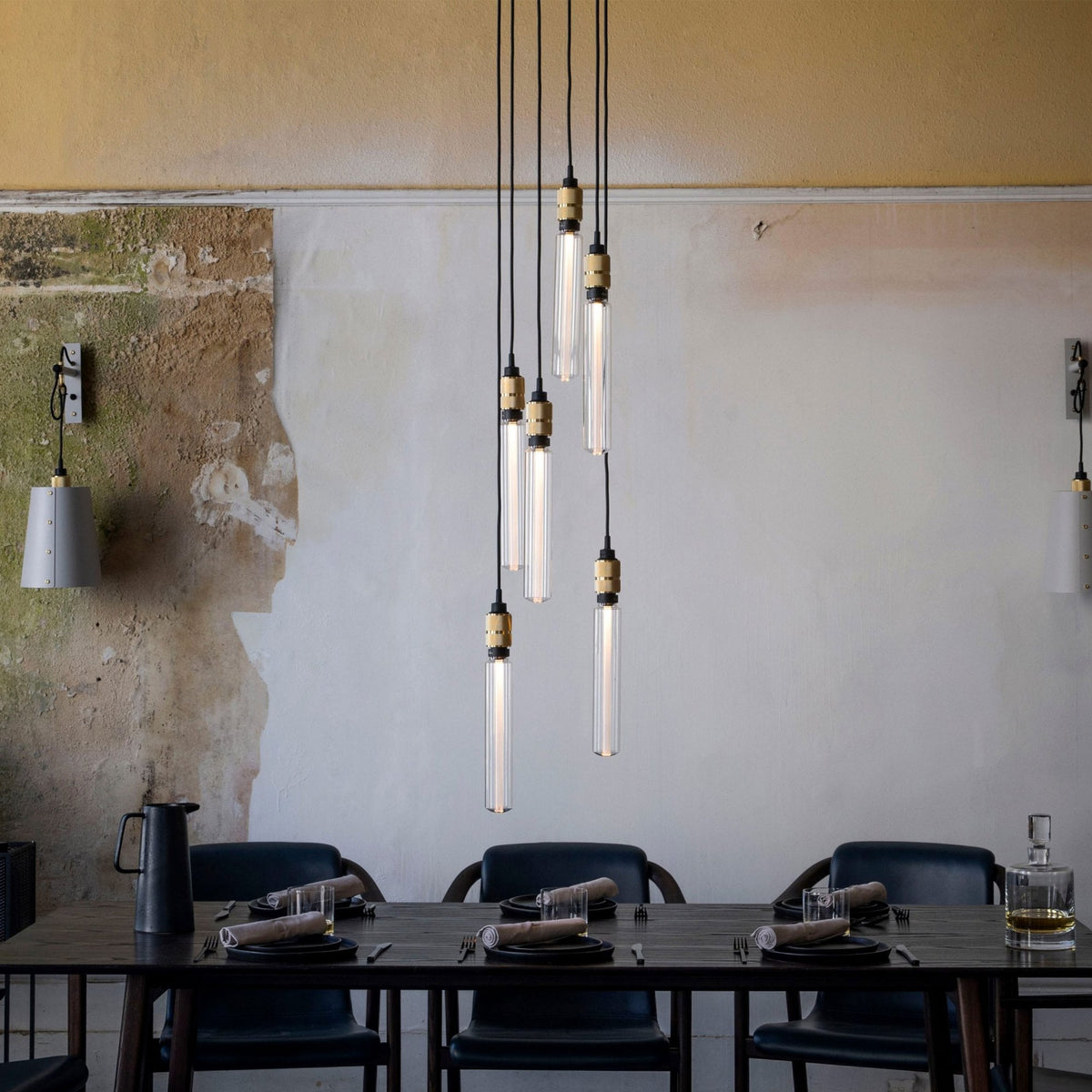A dining room with a long dark wooden table set with black plates and light-colored napkins. Above the table, several pendant lights with clear glass shades hang at varying heights. The walls are textured, with sections of exposed plaster and painted areas in yellow and white. A black pitcher and a glass decanter are also on the table.