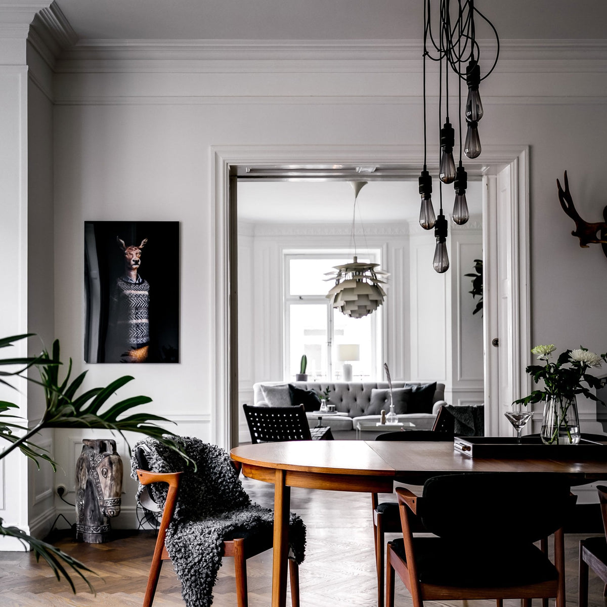 A dining room with a wooden table and chairs, a deer portrait, and a view into a living room.