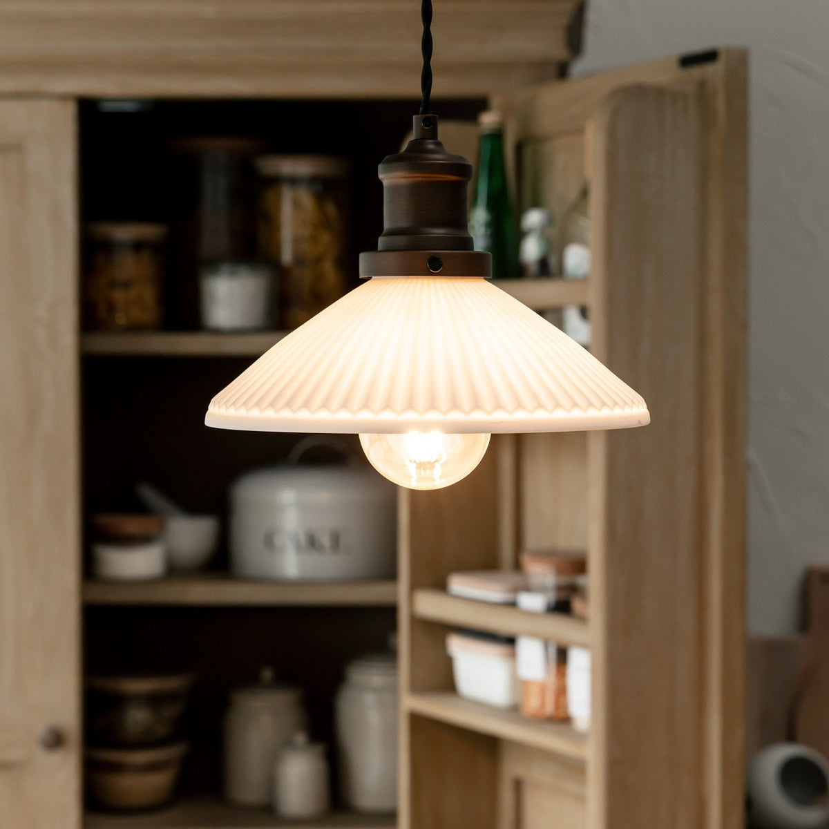 A close-up of a white pleated pendant light with a warm glowing bulb, hanging in front of a wooden cabinet.
