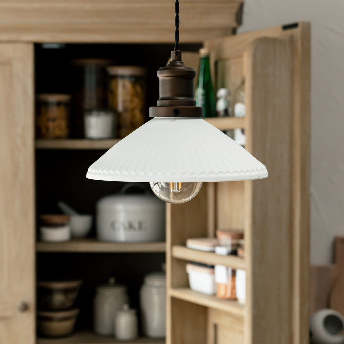 A white fluted glass pendant light with an exposed bulb hangs in front of an open wooden cabinet.