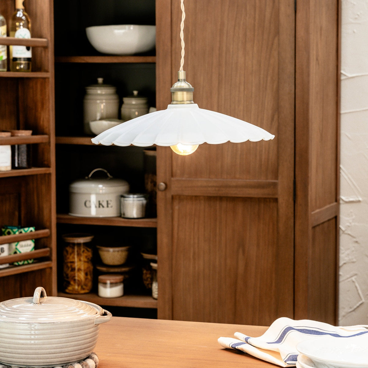 A white scalloped pendant light hangs over a wooden table with a pot and striped napkin.