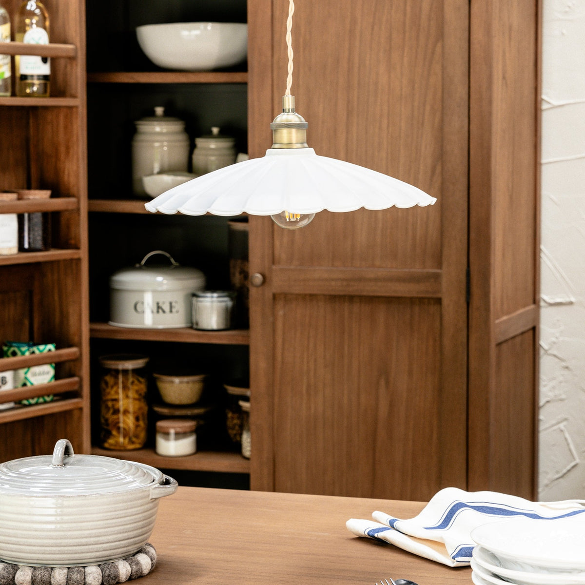 A white scalloped pendant light hangs over a wooden table with a pot, dishes, and a striped napkin.