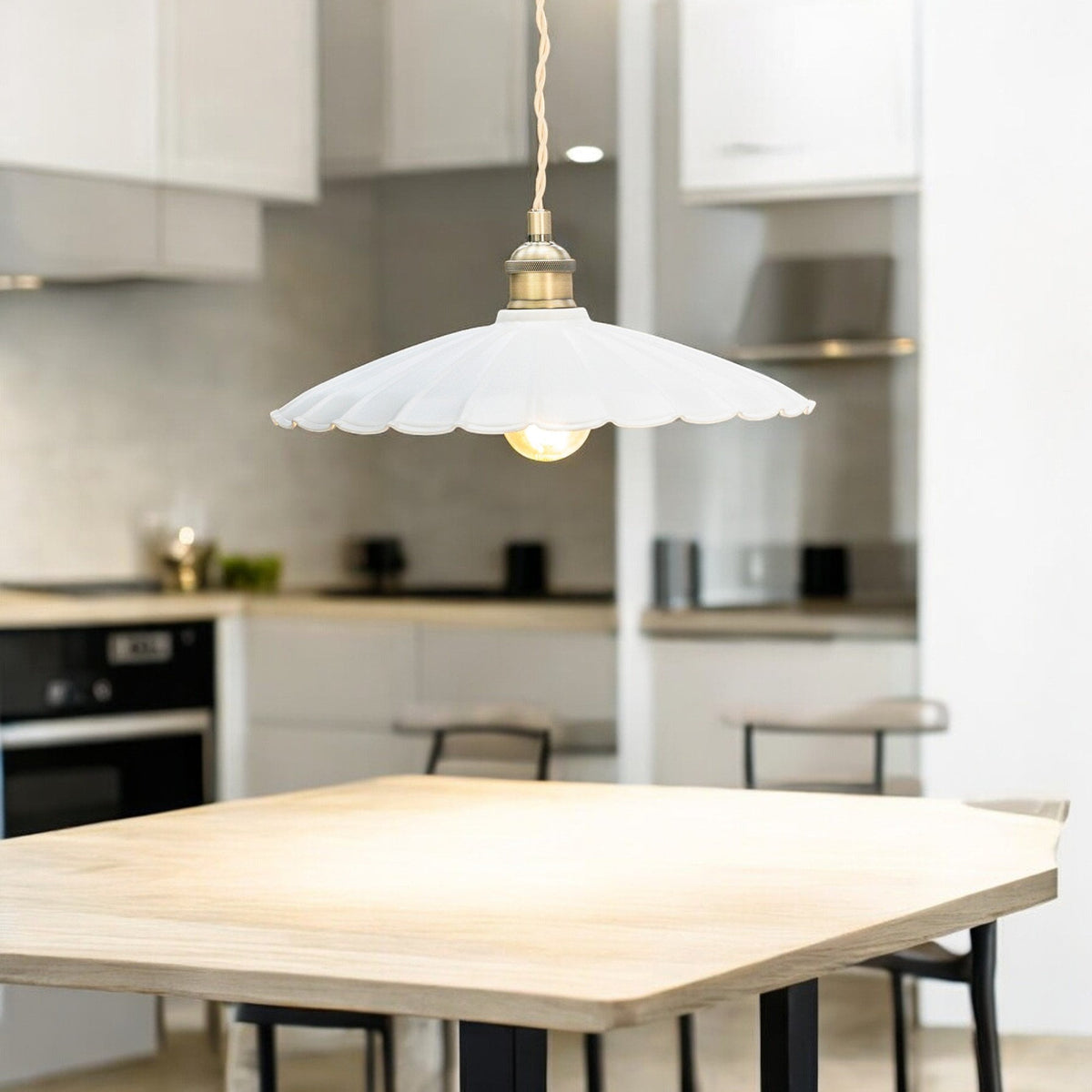 A white scalloped pendant light with a brass fitting hangs over a light wood table in a modern kitchen.