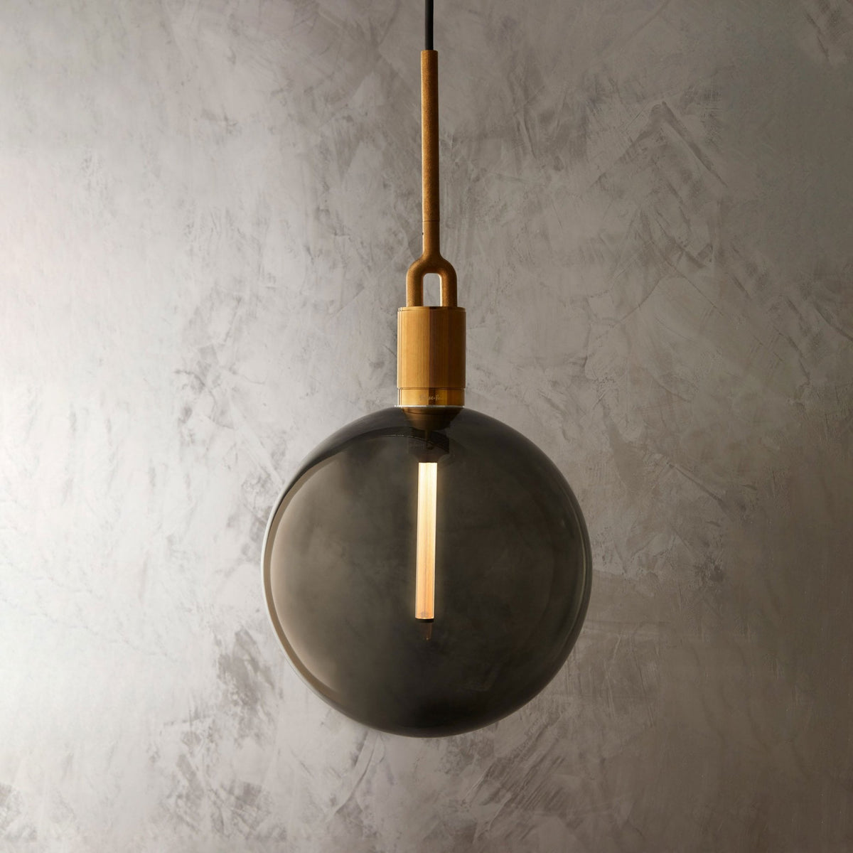 A modern pendant light with a smoked glass globe and a brass fitting, hanging against a textured grey wall.