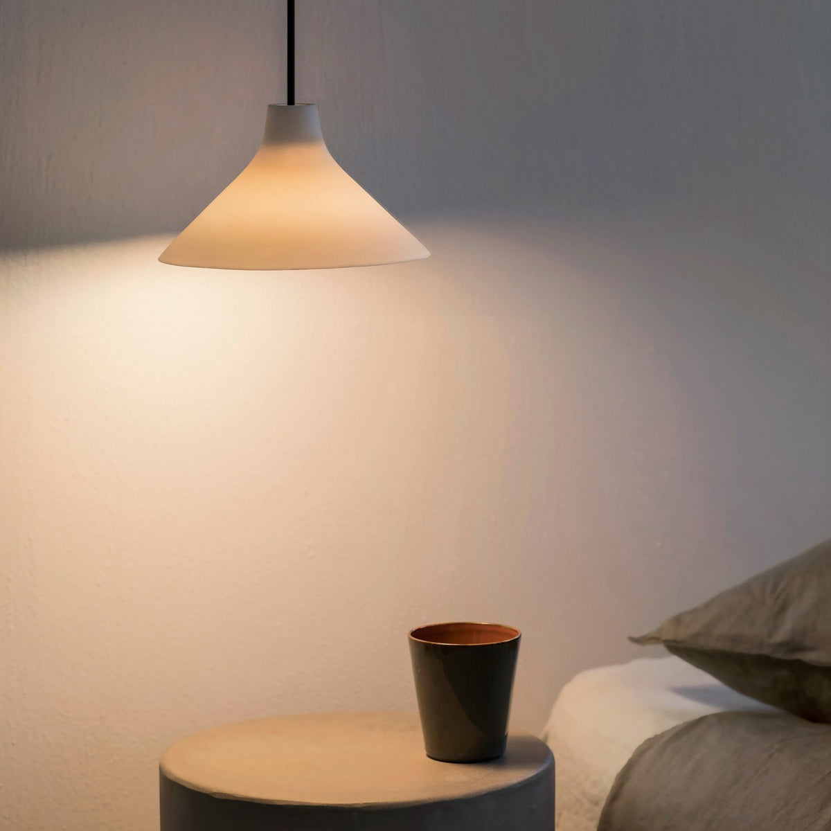 A minimalist white pendant light illuminates a bedside table with a cup.