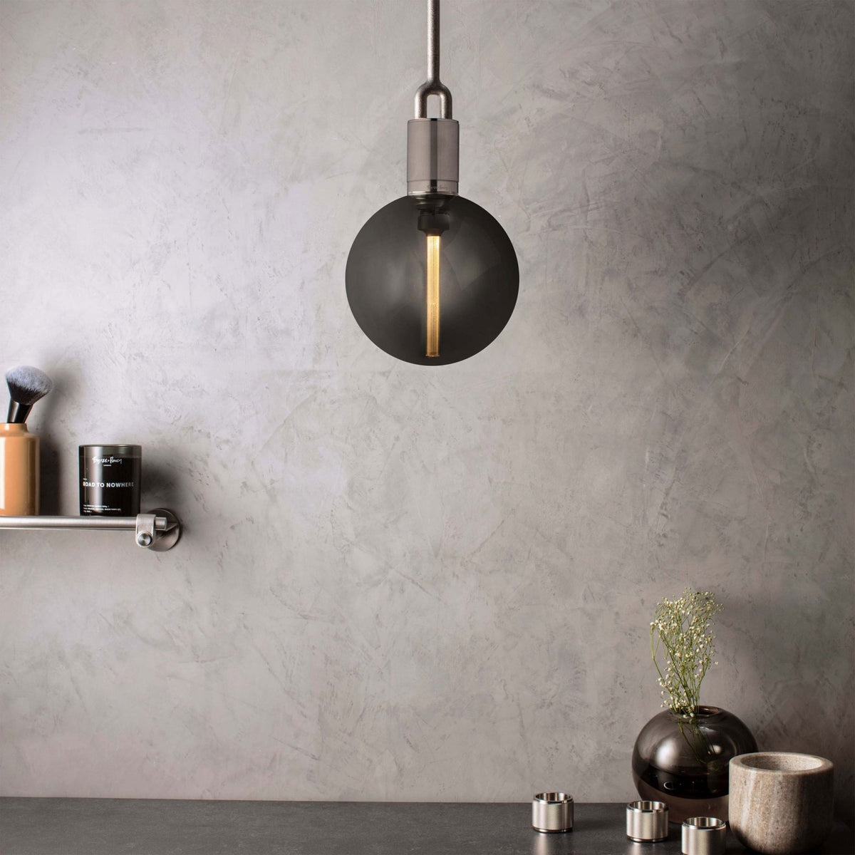 A modern gray light bulb hangs from the ceiling against a textured gray wall, with a shelf and decor below.