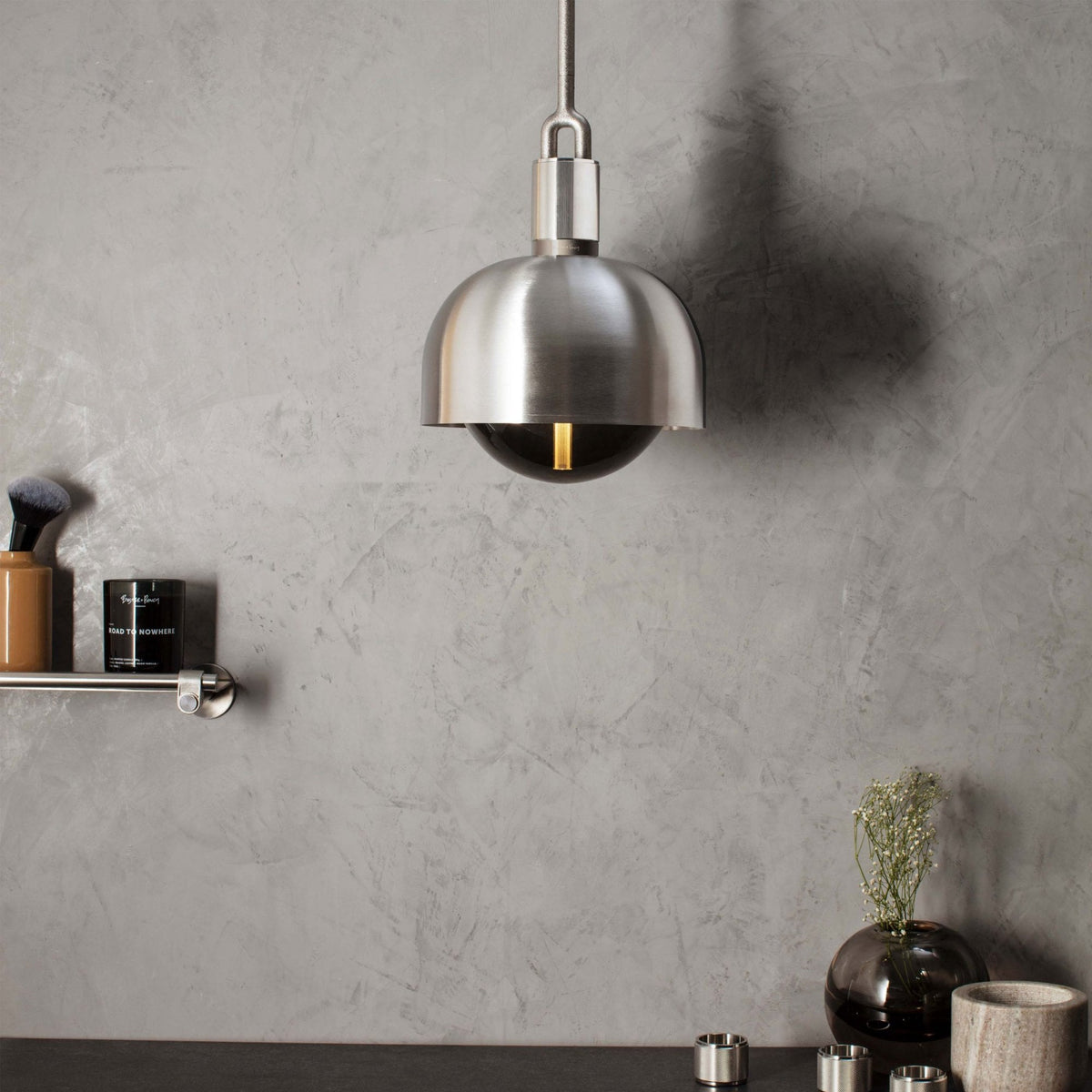A modern brushed metal pendant light hangs over a dark countertop with decorative items against a grey wall.