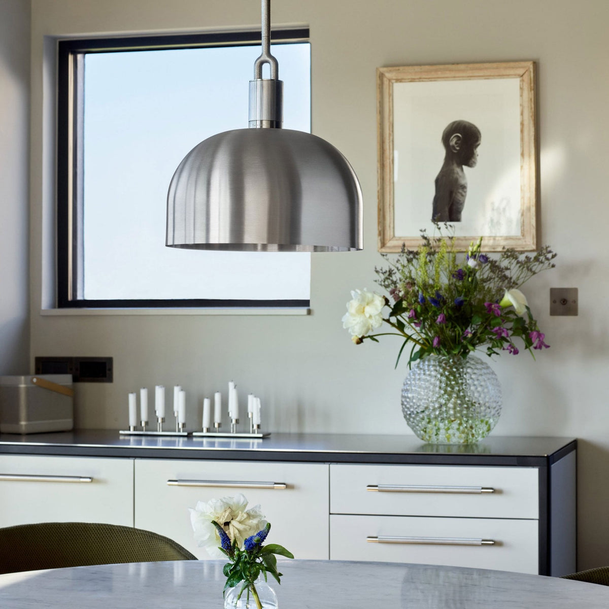 A modern kitchen with a large window, a brushed steel pendant light, and a vase of flowers.