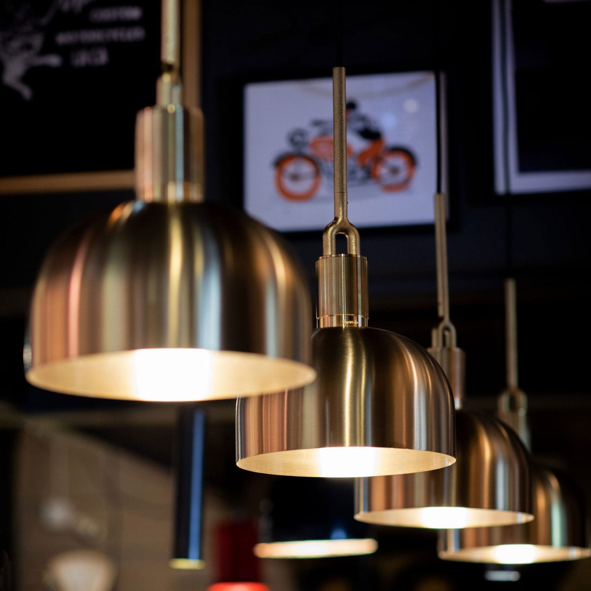 Four brass pendant lights illuminate a dark room, with a blurred orange motorcycle print in the background. 