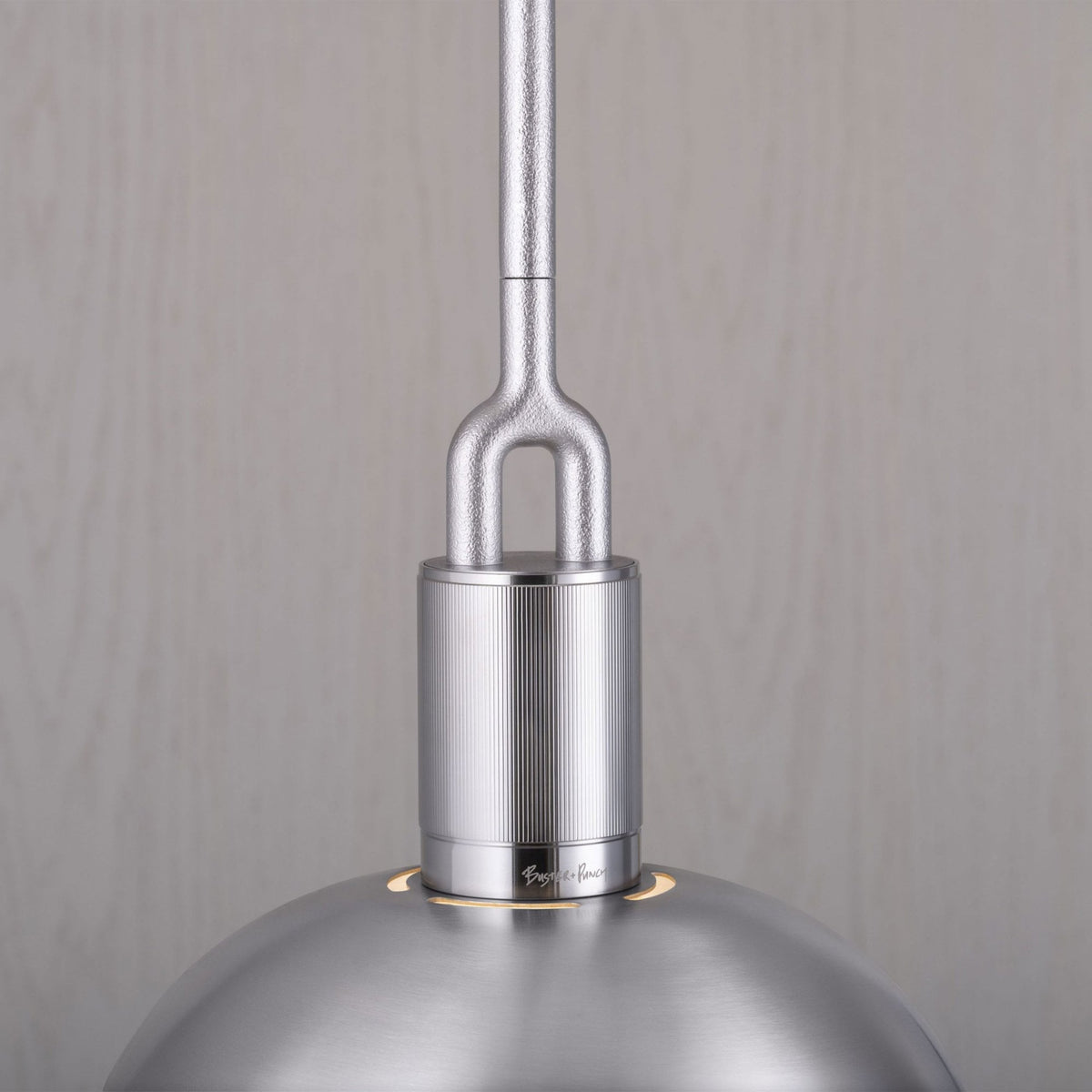 Close-up of a silver pendant light with a textured rod and a ribbed cylindrical base. The light has a smooth, domed shade with small cutouts that emit a warm glow. The brand name Buster + Punch is subtly engraved on the base.