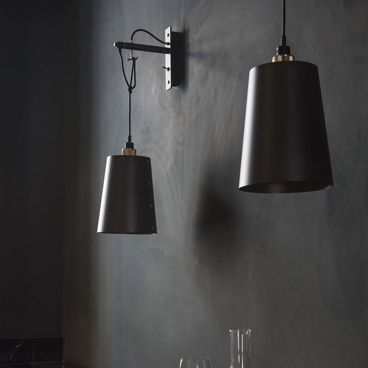 Two black pendant lights with brass accents hang against a dark gray wall, illuminating a table with glassware.