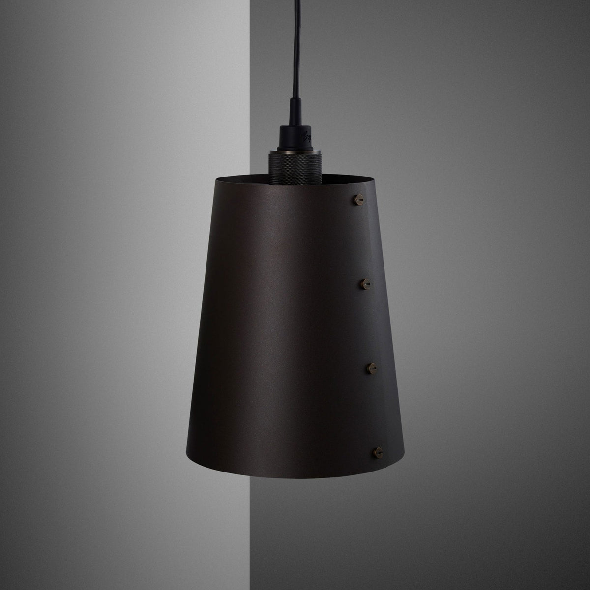 A dark, conical pendant light with visible brass screws, hanging against a grey and white wall.