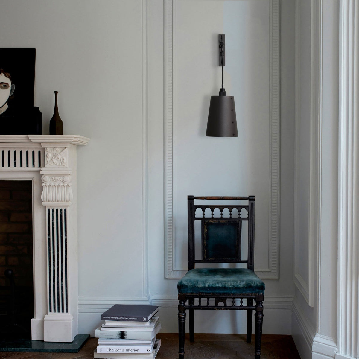 A black wall light with a conical shade hangs above a dark wooden chair with a teal velvet seat.