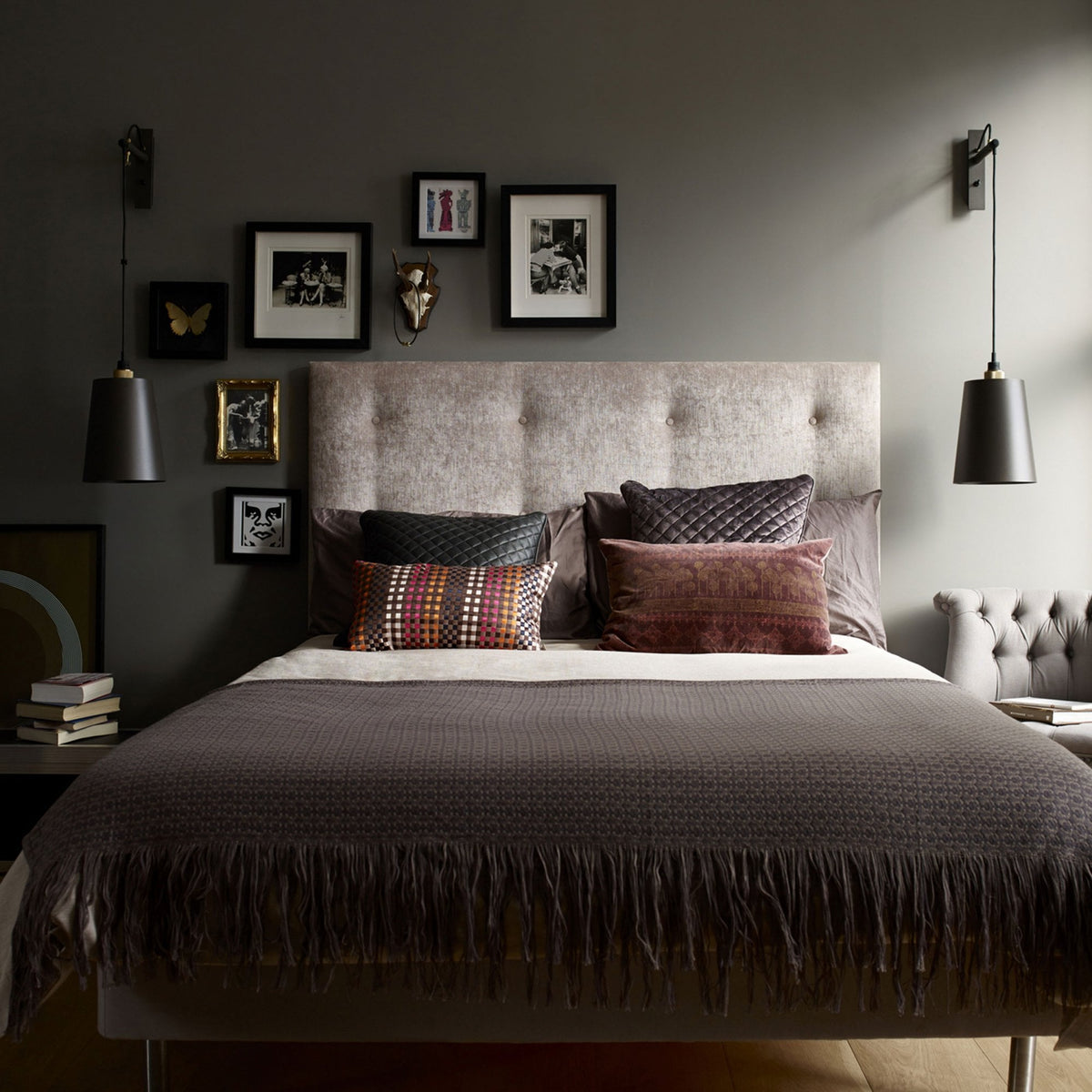 A bedroom with a tufted headboard, dark grey walls, and framed art. A bed with a dark brown throw and pillows.