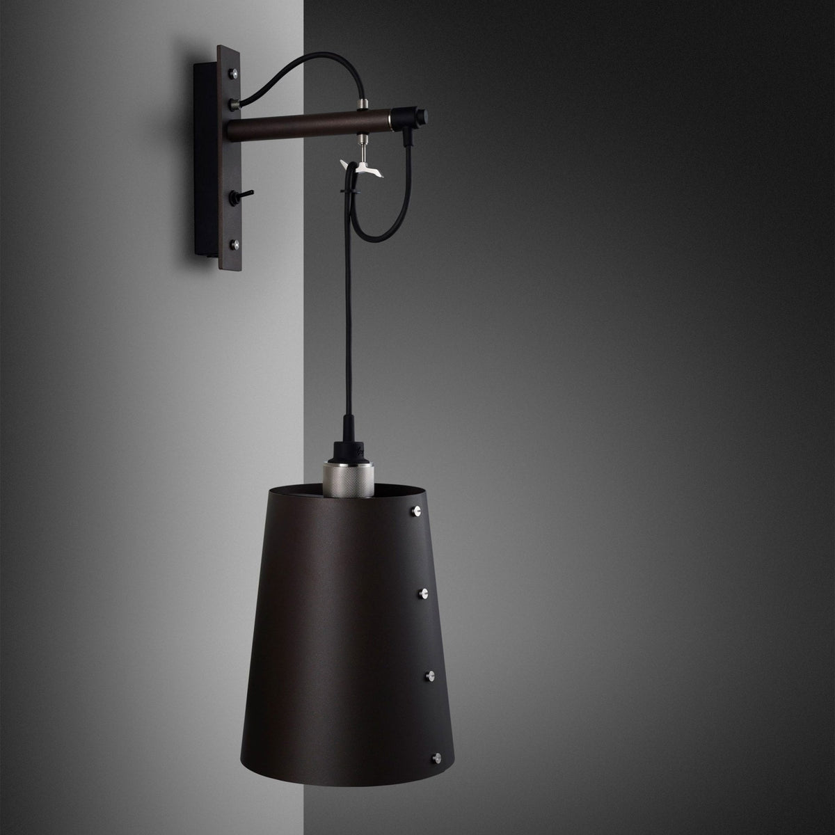 Dark brown wall sconce with a conical shade and exposed silver rivets, mounted on a grey wall.