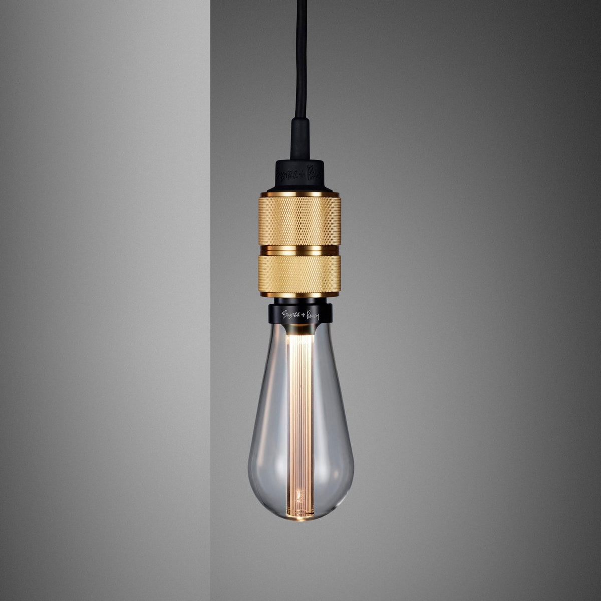 A clear glass light bulb with a gold textured base hangs from a black cord against a gray background.