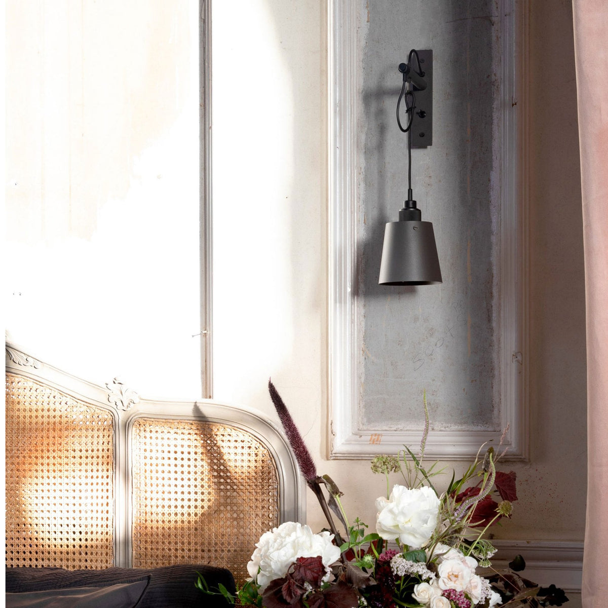 A dark grey wall lamp hangs above a bouquet of white and burgundy flowers, next to a rattan headboard.