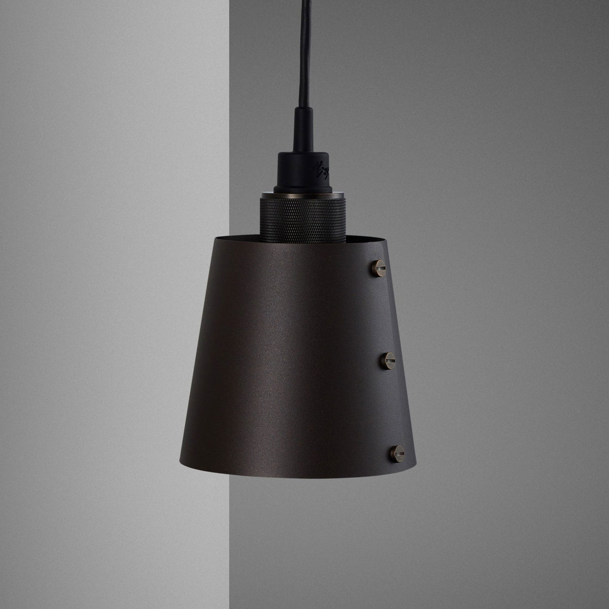 A dark, conical pendant light with visible brass screws, hanging against a grey wall.