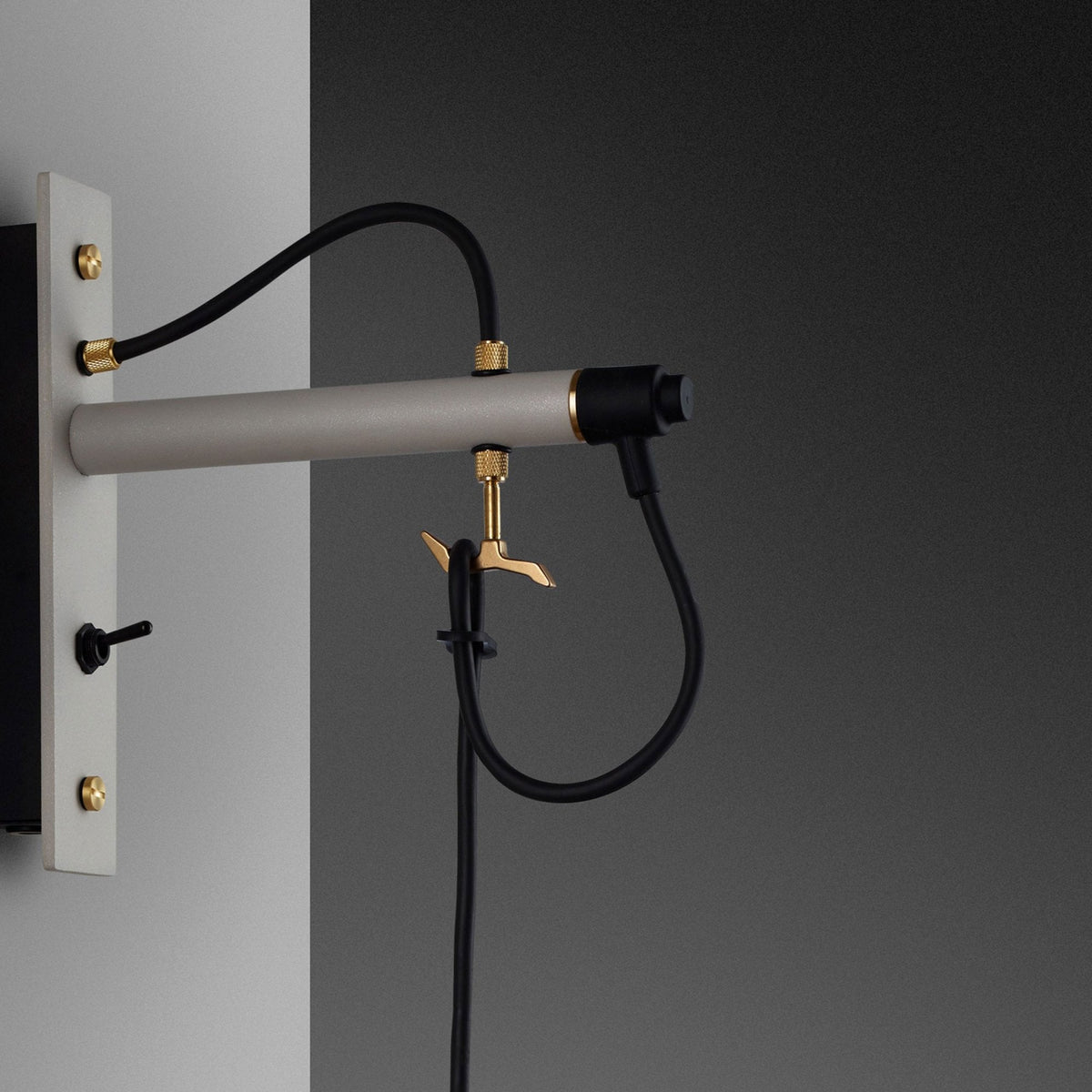 Close-up of a wall-mounted light fixture with a grey bar, black cables, and brass accents. 