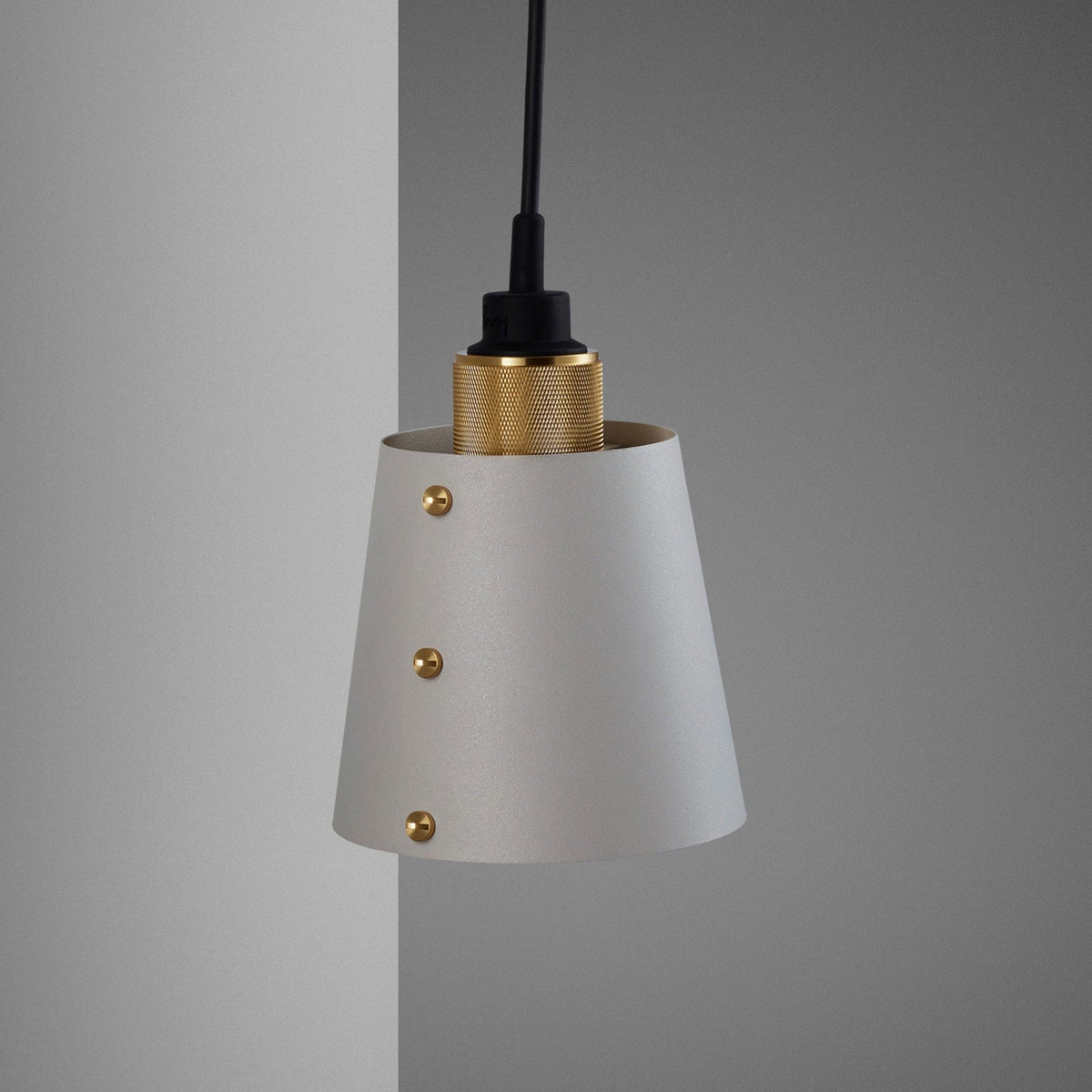 A modern pendant light with a light grey conical shade and a brass-textured top section. It hangs against a grey wall.