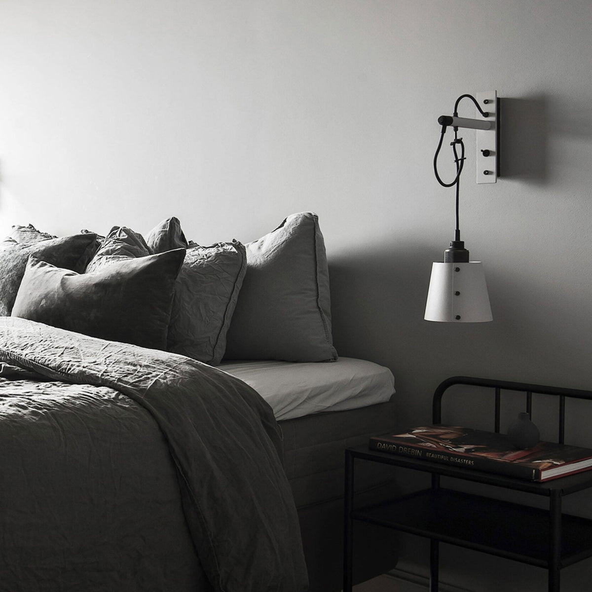 A cozy bedroom with a bed made with dark grey linen bedding and pillows. A black metal nightstand with a book and a small vase sits next to the bed. A white wall-mounted lamp hangs above the nightstand.