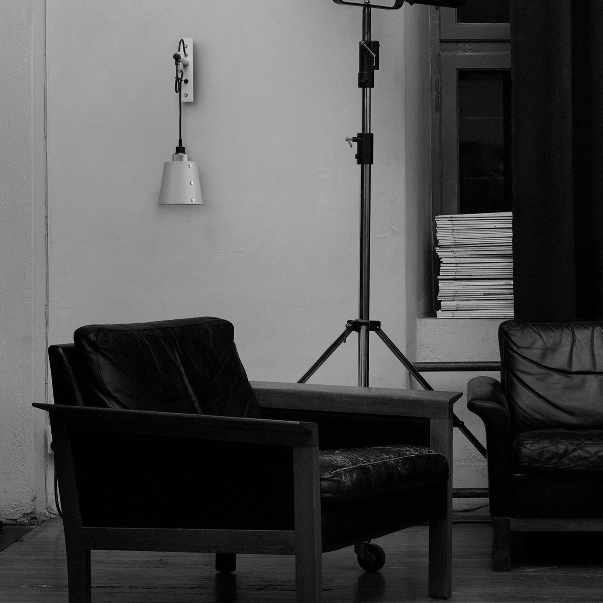 Black and white image of two leather armchairs and a tripod light stand in a room with a wall-mounted lamp.
