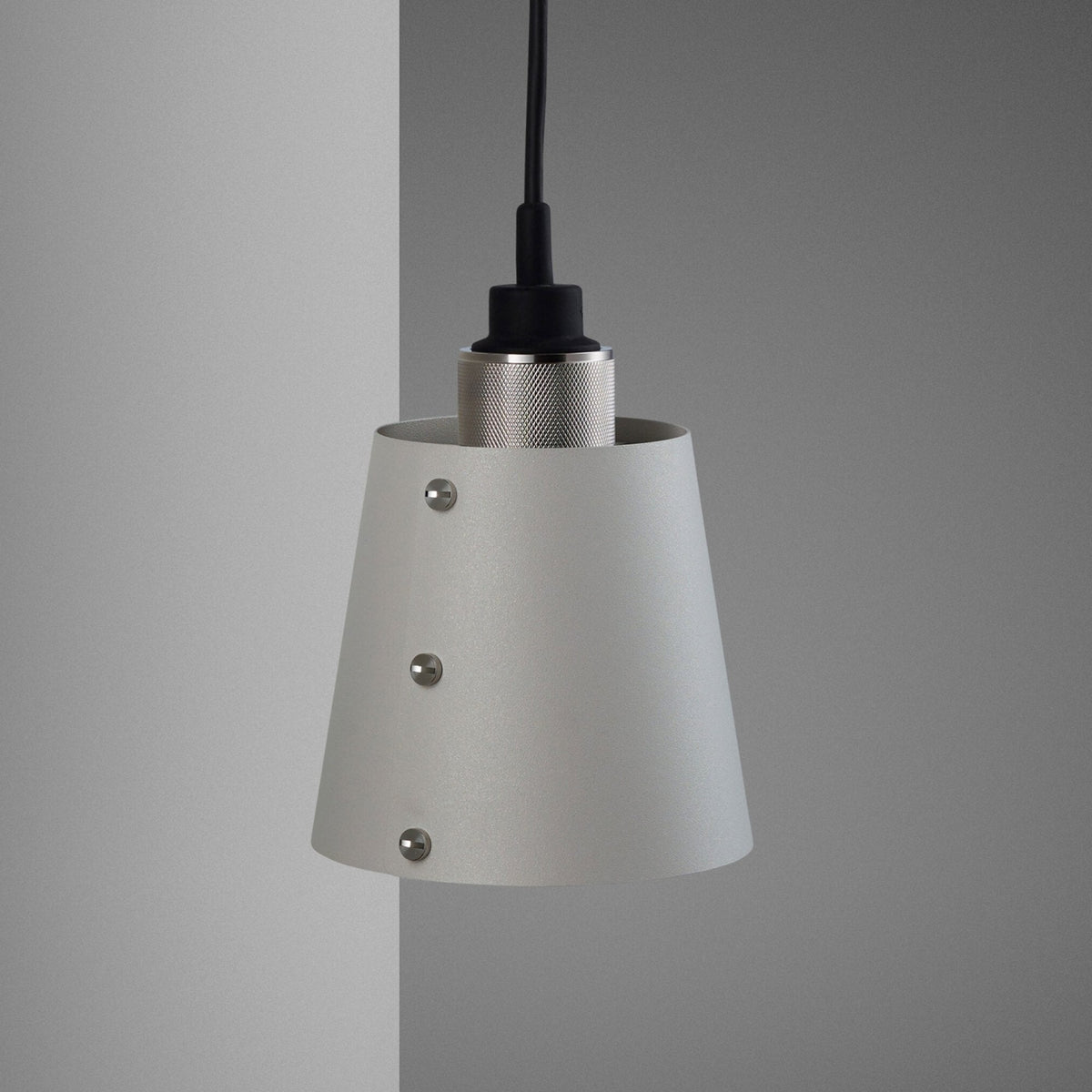 A close-up of a light grey conical pendant light with three visible silver screws, hanging against a grey wall.