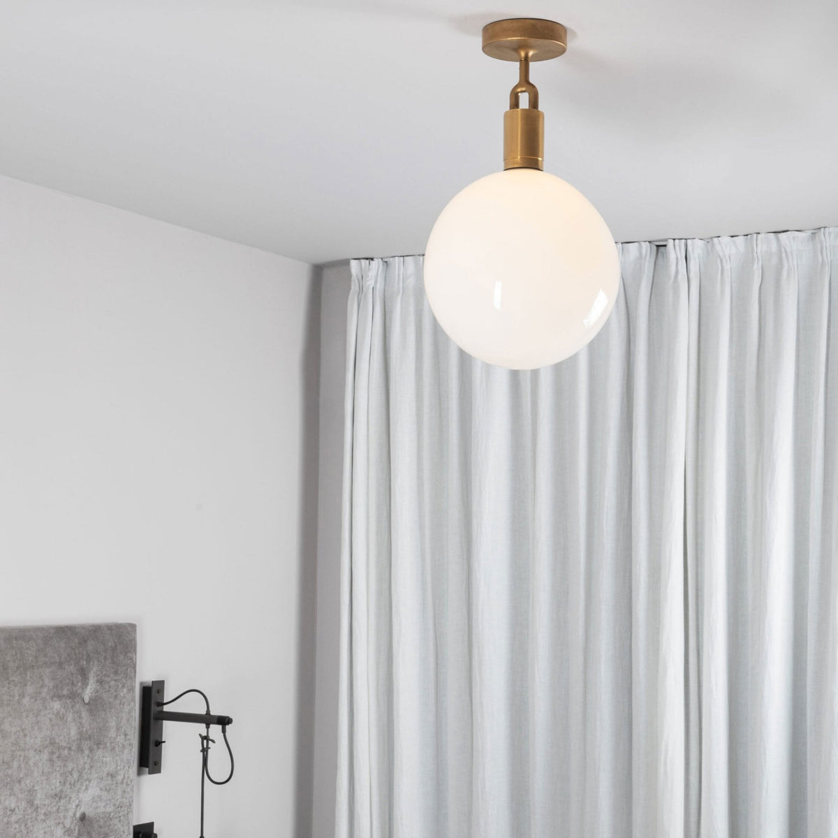 A modern brass ceiling light with a white globe shade illuminates a room with light grey walls and white curtains.