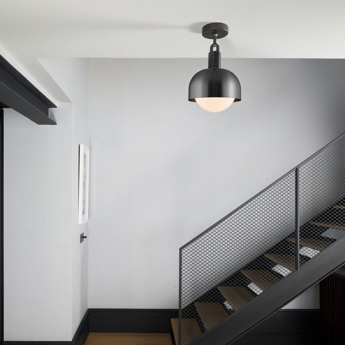 A modern black semi-flush mount light fixture illuminates a staircase with a black metal mesh railing.