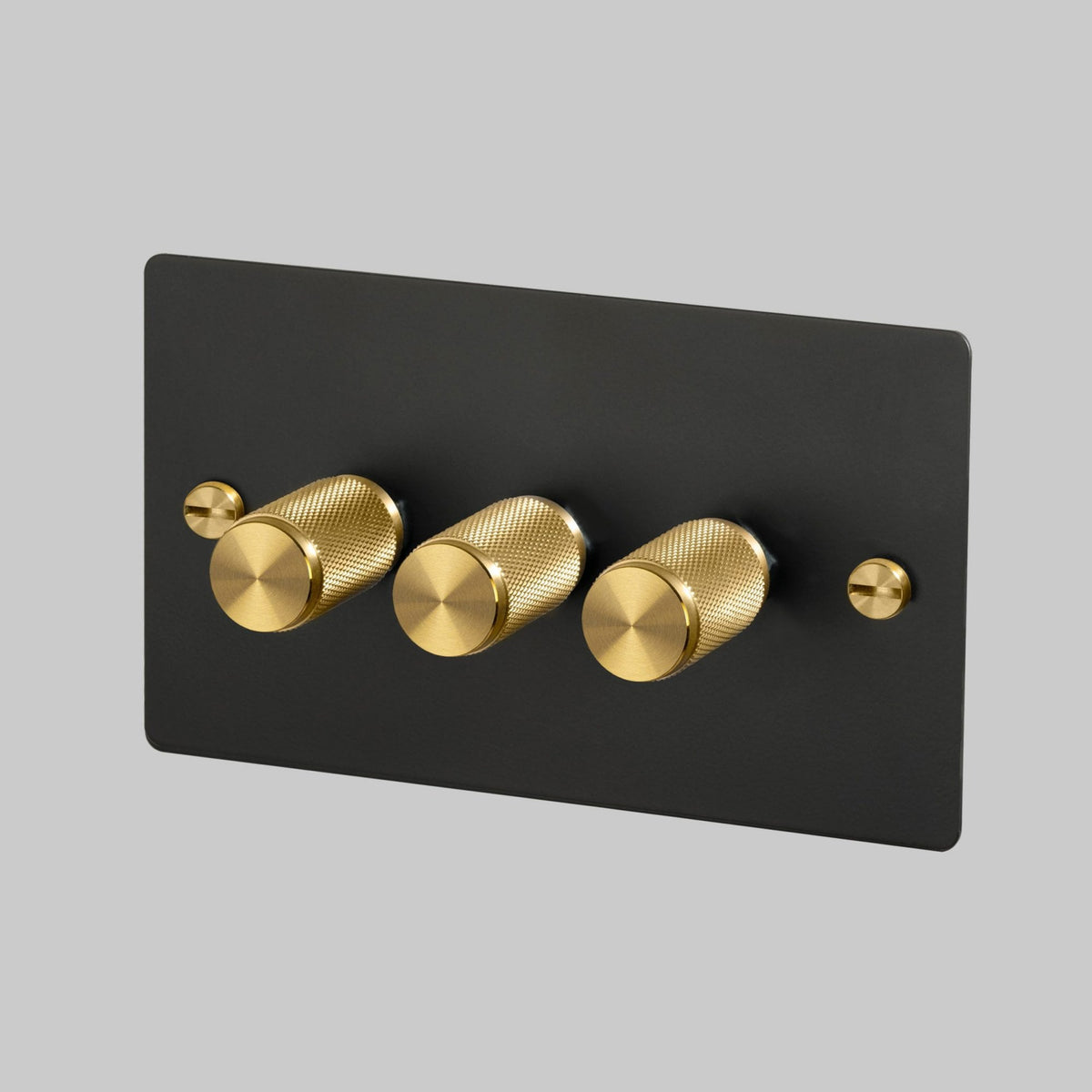 A sleek black rectangular wall plate features three cylindrical brass dimmer knobs and brass screws.