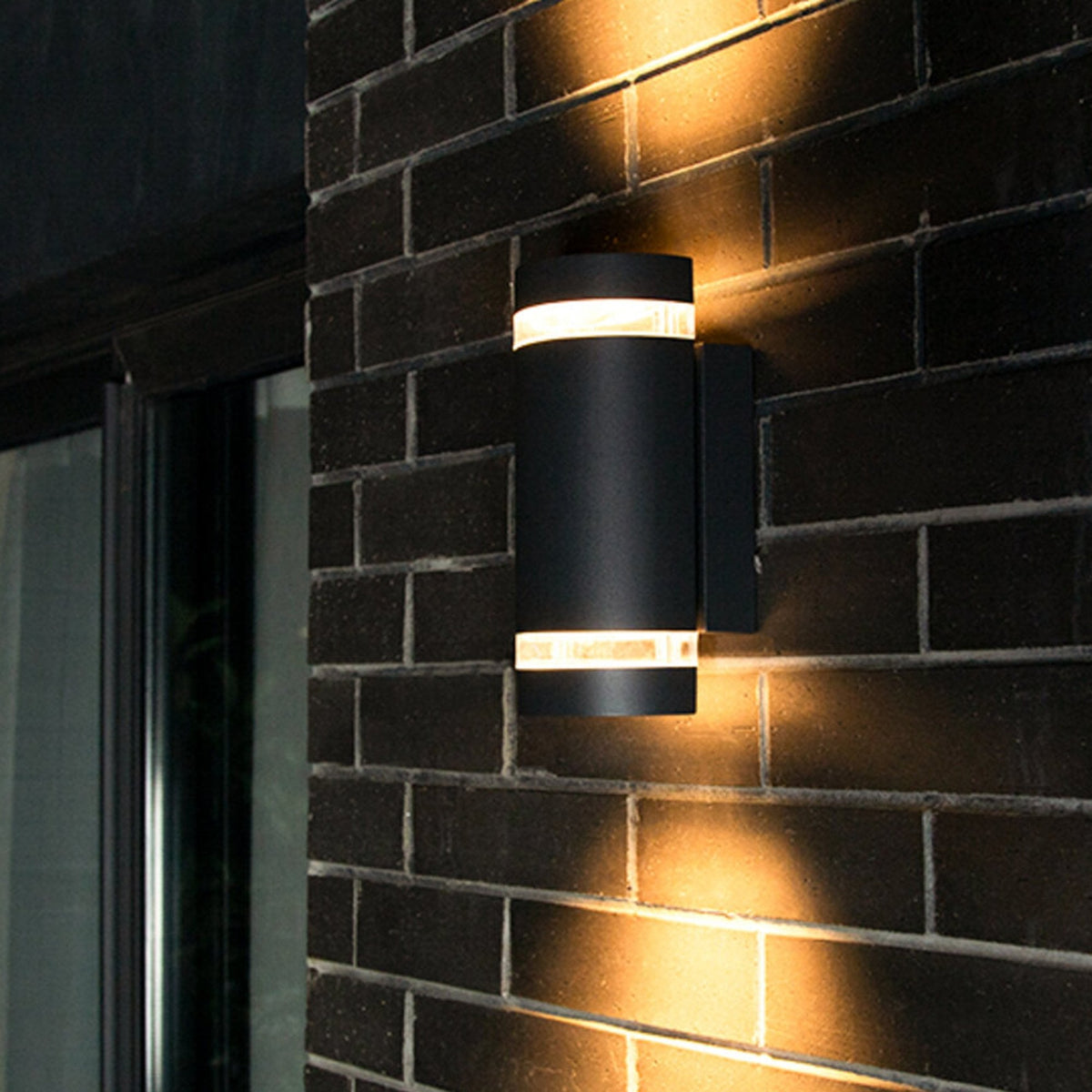 Black cylindrical outdoor wall light with two illuminated bands, casting warm light on a dark brick wall.