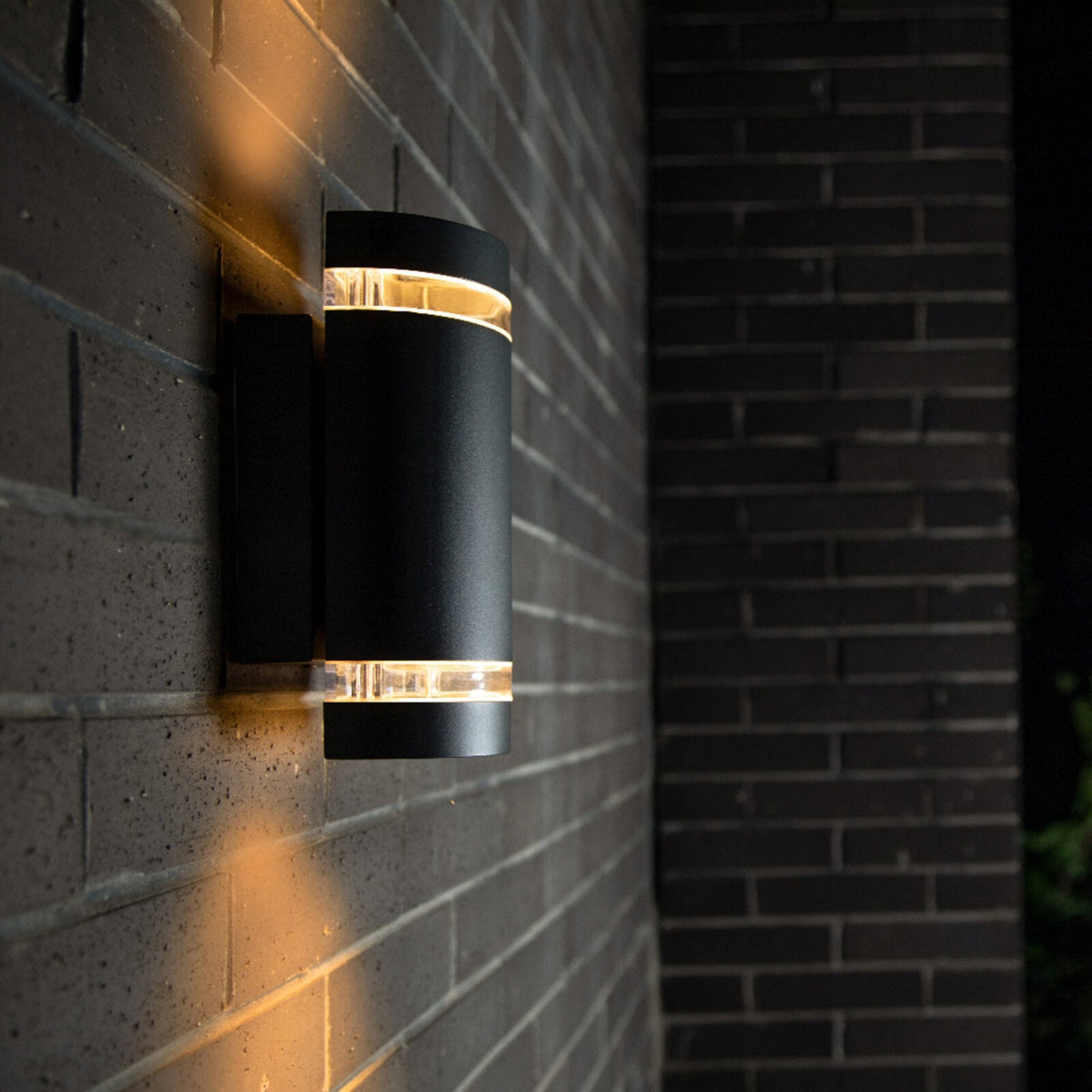 Modern black cylindrical outdoor wall light with warm glow on a dark brick wall at night.