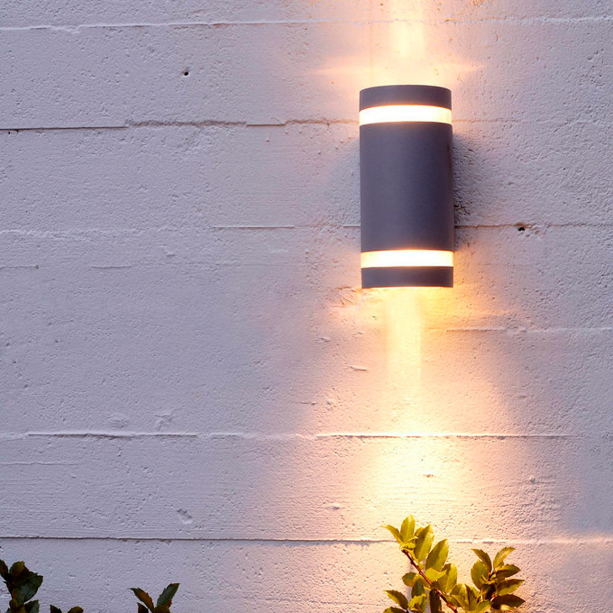 Modern grey cylindrical outdoor wall light illuminating a textured white wall and green foliage.
