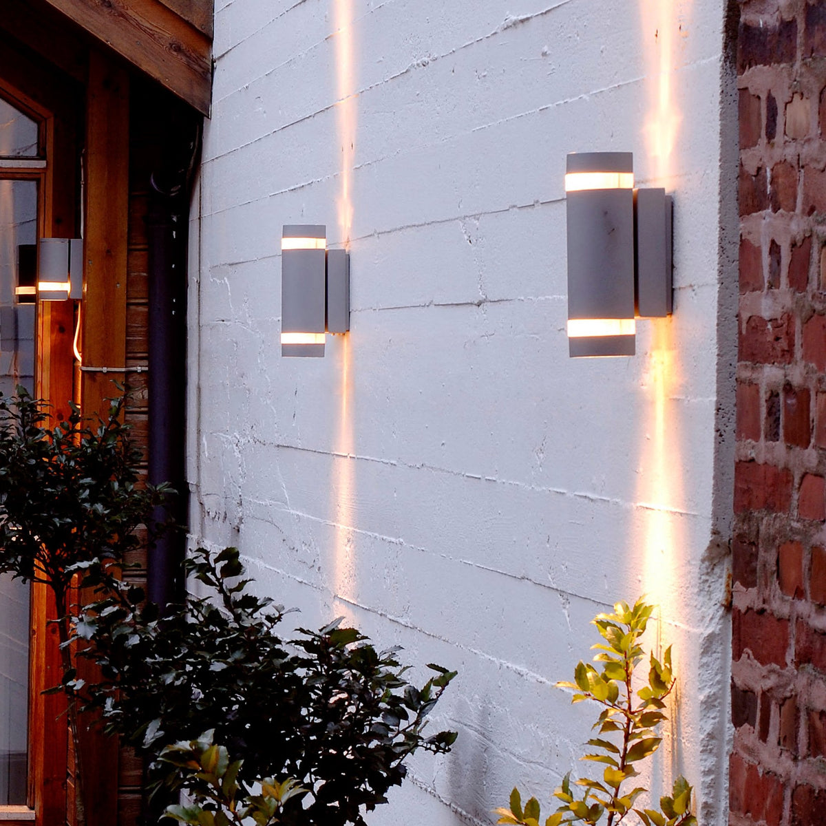 Two up-down wall lights illuminate a grey wall at night, casting light upwards and downwards.