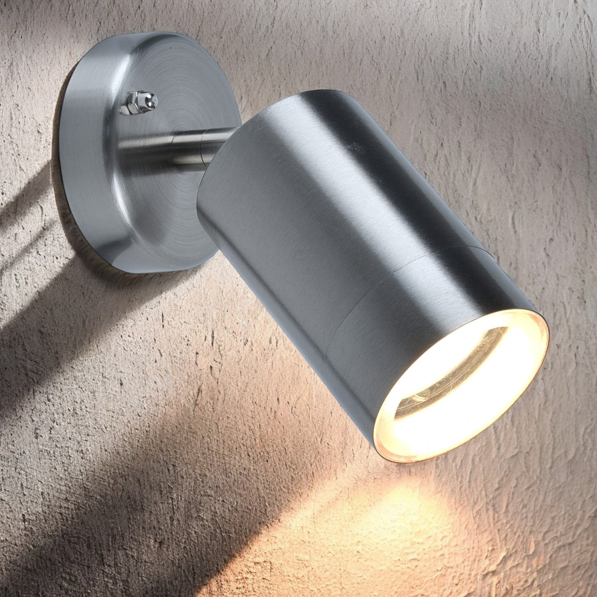Brushed metal wall-mounted spotlight illuminates a textured wall with warm light, casting shadows.