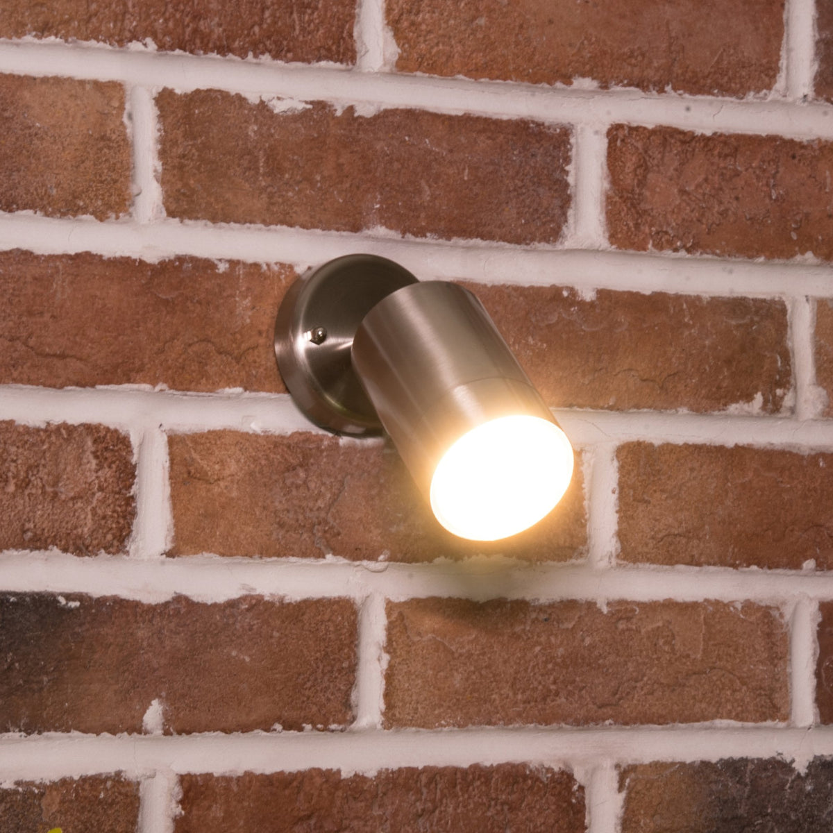 A close-up of a brushed metal spotlight with a warm glow, mounted on a rustic brick wall.