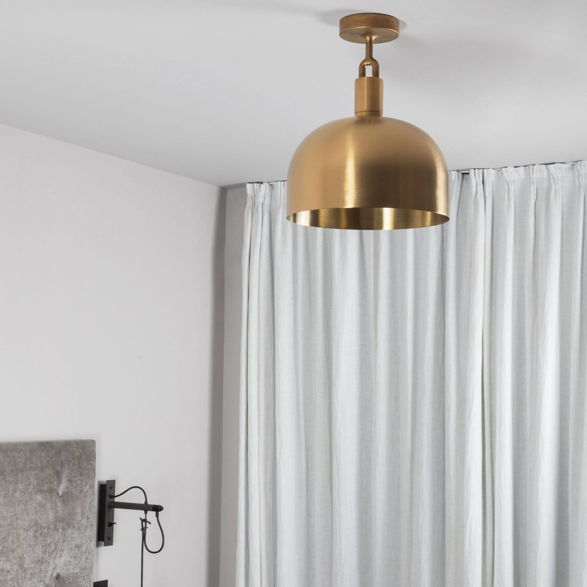A gold dome-shaped ceiling light hangs above a grey upholstered headboard and white curtains.