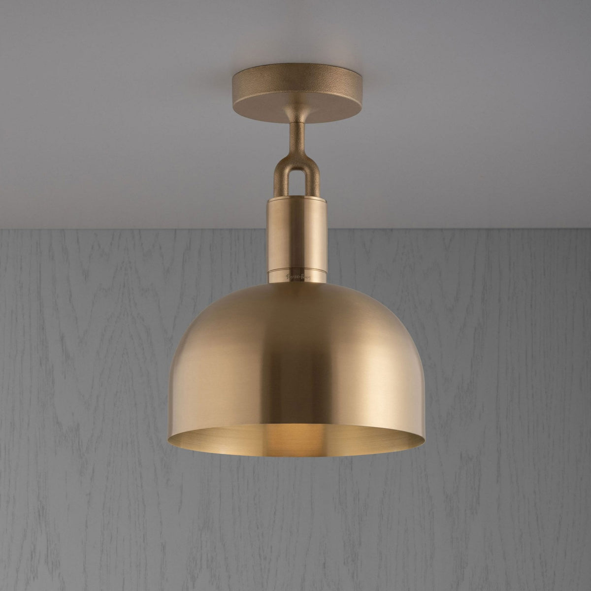 A brass ceiling light with a domed shade hangs from a white ceiling against a grey wooden wall.